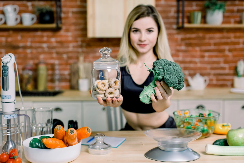 Maya Thompson in her kitchen, holding a head of broccoli in one hand and a jar of donuts in the other, contemplating a healthy choice.