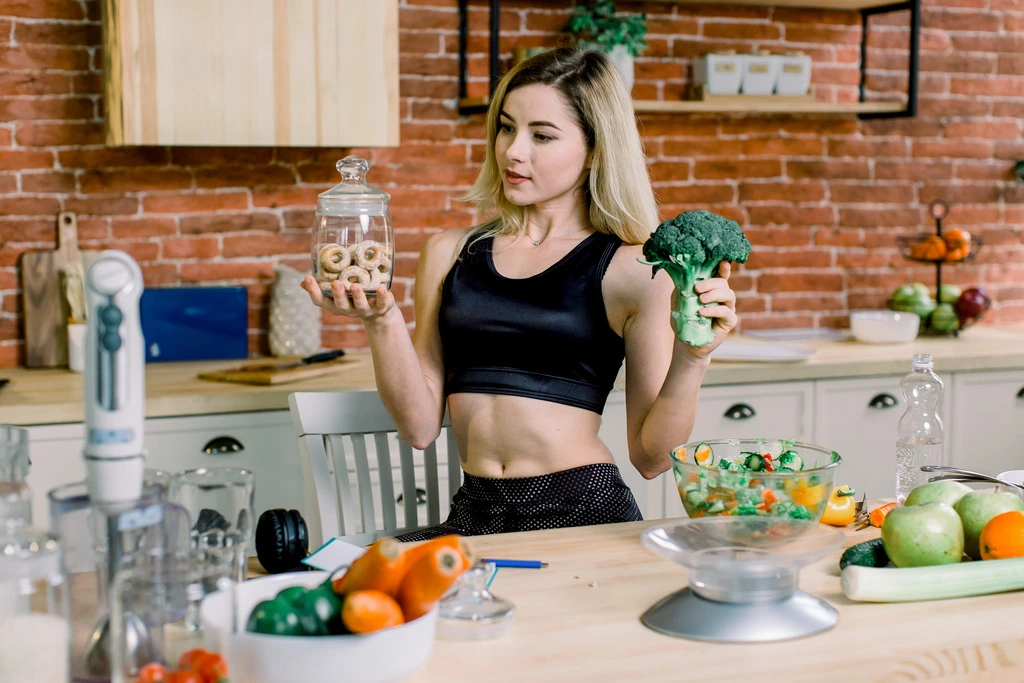 Maya Thompson holds up healthy broccoli and a jar of donuts, smiling to show her balanced and realistic approach to nutrition.