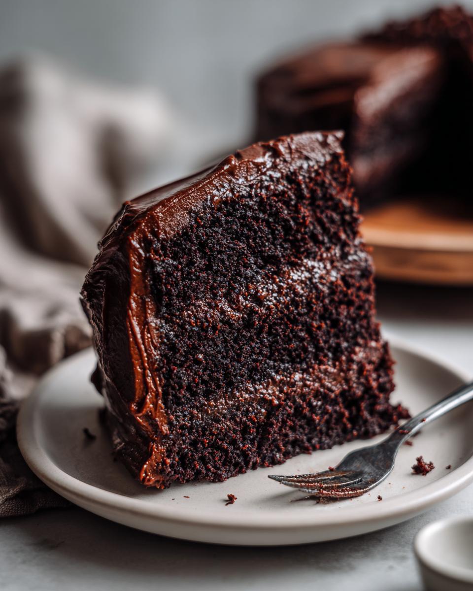 A rich, moist slice of chocolate cake covered in glossy frosting, served on a small plate with a fork.