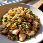 A close-up, appetizing shot of homemade chicken fried rice mixed with bright green peas and scrambled egg in a white bowl.