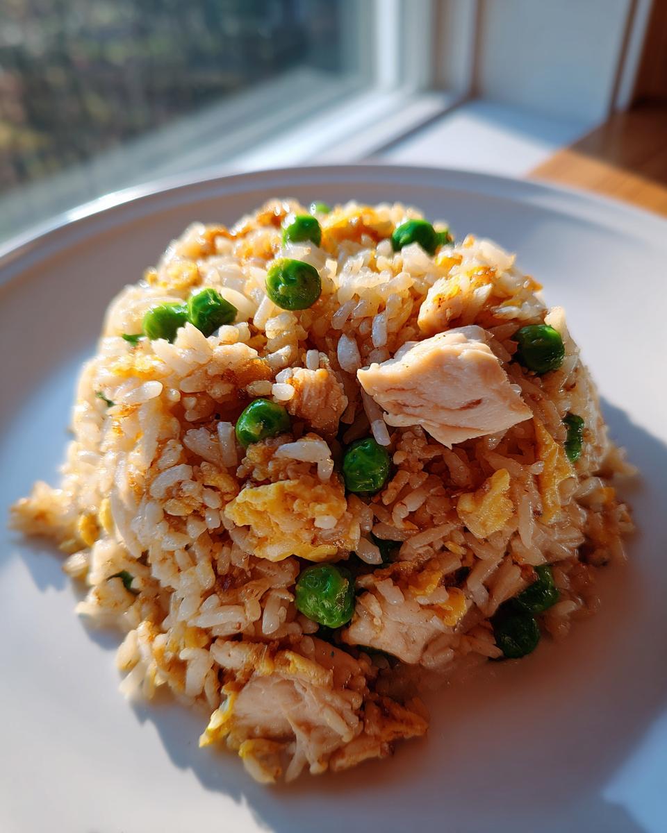 A mound of homemade chicken fried rice featuring chunks of chicken, scrambled egg, and bright green peas, served on a white plate.