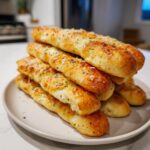 A tall stack of golden brown, cheesy garlic breadsticks topped with herbs on a light plate.