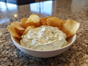 A white bowl filled with creamy, herbed pickle dip, surrounded by ruffled potato chips.