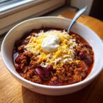 A close-up, appetizing bowl of rich crockpot chili topped with shredded cheese and a dollop of sour cream.