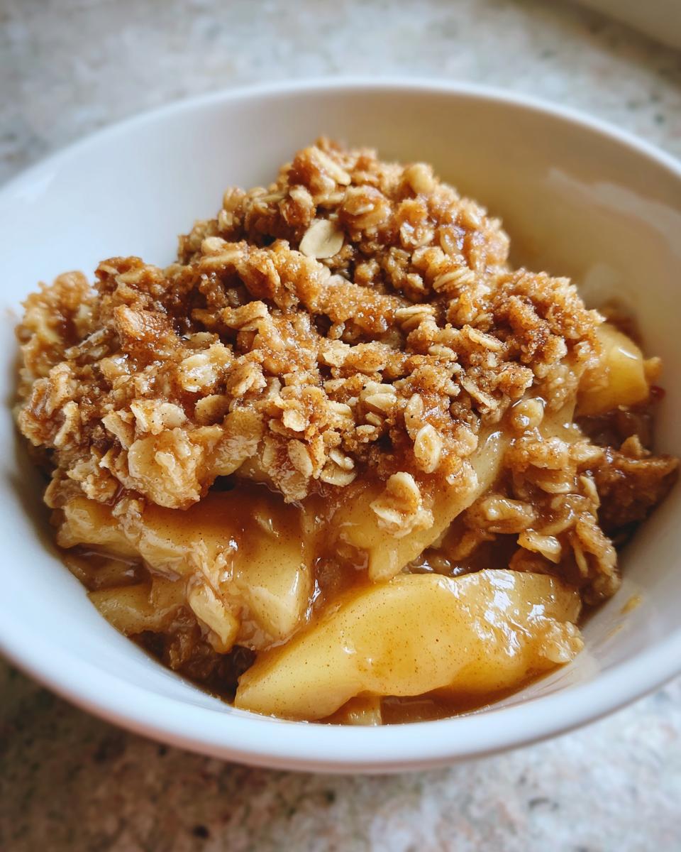 A close-up of a single serving of warm apple crisp recipe in a white bowl, showing soft spiced apples under a crunchy oat topping.