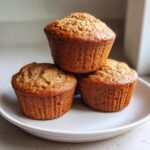Three golden-brown banana protein muffins stacked on a light gray plate, ready to eat.
