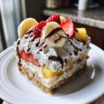 A generous slice of banana split cake featuring a graham cracker crust, creamy filling, and topped with whipped cream, strawberries, bananas, and chocolate drizzle.