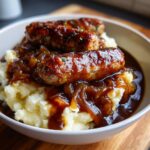 A close-up shot of three glazed sausages served over creamy mashed potatoes, covered in rich onion gravy, a classic bangers and mash dish.