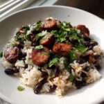 A white bowl filled with white rice, black beans and rice, topped with sliced, grilled sausage and fresh cilantro.