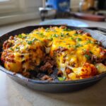 Close-up of Cheesy Taco Potatoes baked in a cast-iron skillet, topped with melted cheddar and green onions.