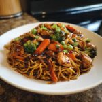 A close-up of a plate piled high with delicious chicken chow mein recipe, featuring noodles, chicken pieces, carrots, and broccoli.