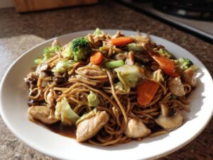 A close-up of a plate piled high with homemade chicken chow mein recipe, featuring noodles, chicken pieces, broccoli, carrots, and mushrooms.