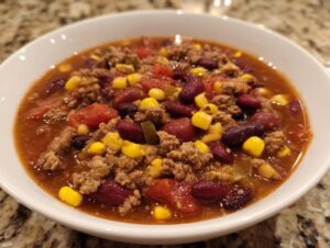 Close-up of a white bowl filled with rich, savory cowboy soup featuring ground beef, kidney beans, corn, and tomatoes.