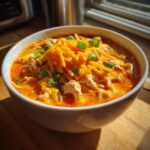 Close-up of a bowl of creamy buffalo chicken chili, topped with shredded cheddar cheese and green onions.
