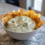 A white bowl filled with creamy pickle dip, garnished with dill, and surrounded by crispy potato chips.