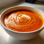 Close-up of a vibrant orange, creamy tomato bisque in a white bowl, garnished with a swirl of cream and black pepper.