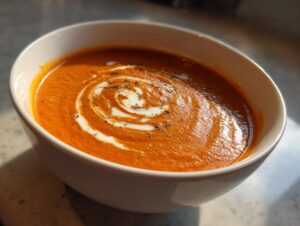 Close-up of a vibrant orange bowl of creamy tomato bisque, swirled with cream and sprinkled with pepper.