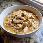 Close-up of a white bowl filled with creamy crockpot white chicken chili, featuring shredded chicken and white beans.