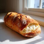 A golden-brown, crusty loaf of homemade french bread resting on a white marble surface near a window.