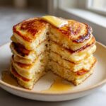 A tall stack of fluffy greek yogurt pancakes, cut to show the interior, topped with melting butter and drizzled with syrup.