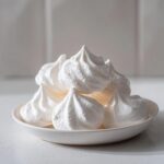 A close-up of several perfectly piped, white meringue cookies stacked in a small white bowl.