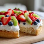 Close-up of a fruit pizza sugar cookie with a slice removed, topped with whipped cream, strawberries, kiwi, and blueberries.
