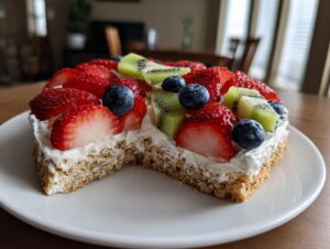 A close-up of a fruit pizza sugar cookie with a bite taken out, topped with cream cheese frosting, strawberries, blueberries, and kiwi.