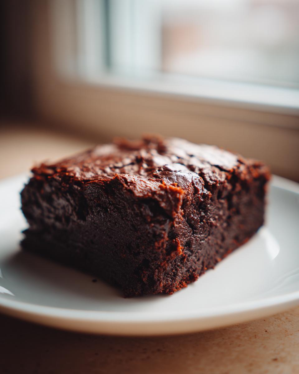 A close-up, dark, and fudgy square of sweet potato brownies resting on a white plate near a window.