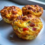 Close-up of three golden brown Egg Muffin Cups filled with visible bits of ham and red pepper on a white plate.