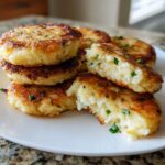 A stack of golden brown, pan-fried mashed potato cakes, one cut open to show the fluffy interior with chives.