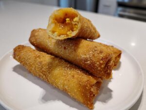 A stack of crispy, golden-brown peach cobbler egg rolls, with one cut open revealing the warm peach filling.