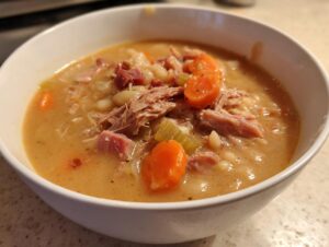 Close-up of a white bowl filled with thick, creamy Ham and Bean Soup, topped with shredded ham, white beans, and sliced carrots.