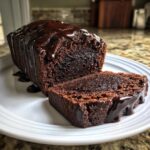 A loaf of rich, dark hot fudge brownie bread with a slice cut, generously drizzled with thick chocolate glaze.