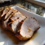 Close-up of several thick slices of juicy pork loin roast with a flavorful herb crust resting in pan juices.