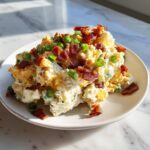 A serving of creamy baked potato salad topped generously with crumbled bacon and chopped green onions.