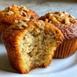 Close-up of a moist banana nut muffin with a bite taken out, revealing the soft crumb and topped with pecans.