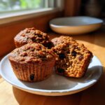 Four moist bran muffins, one cut open showing raisins, served on a white plate in bright sunlight.