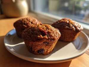 Three golden brown, moist bran muffins resting on a light-colored plate near a sunny window.
