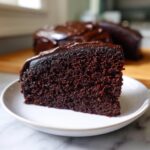 Close-up of a rich, dark slice of chocolate cake with glossy frosting, served on a white plate.