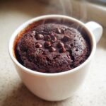 Close-up of a moist, dark chocolate chip mug cake steaming inside a white mug.