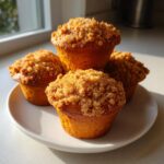 Four moist sweet potato muffins stacked slightly on a white plate, featuring a generous, golden crumb topping.