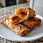 Close-up of crispy, golden peach cobbler egg rolls dusted with cinnamon sugar, one cut open showing the filling.