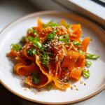 A close-up of a vibrant raw carrot salad made with shaved carrot ribbons, topped with toasted sesame seeds and sliced green onions.