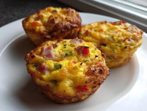 Close-up of three golden-brown Egg Muffin Cups filled with ham and colorful peppers on a white plate.
