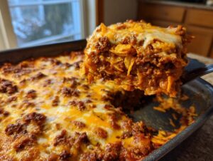 A serving spoon lifts a cheesy, meaty portion of Dorito Casserole out of the baking dish.
