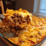 A fork lifts a cheesy, meaty portion of Dorito Casserole out of a glass baking dish.