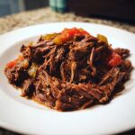 A close-up of tender, shredded Mississippi pot roast served on a white plate, topped with chunks of tomato and peppers.