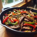 Close-up of sizzling steak fajitas with sliced red and green peppers and onions in a black cast iron skillet.