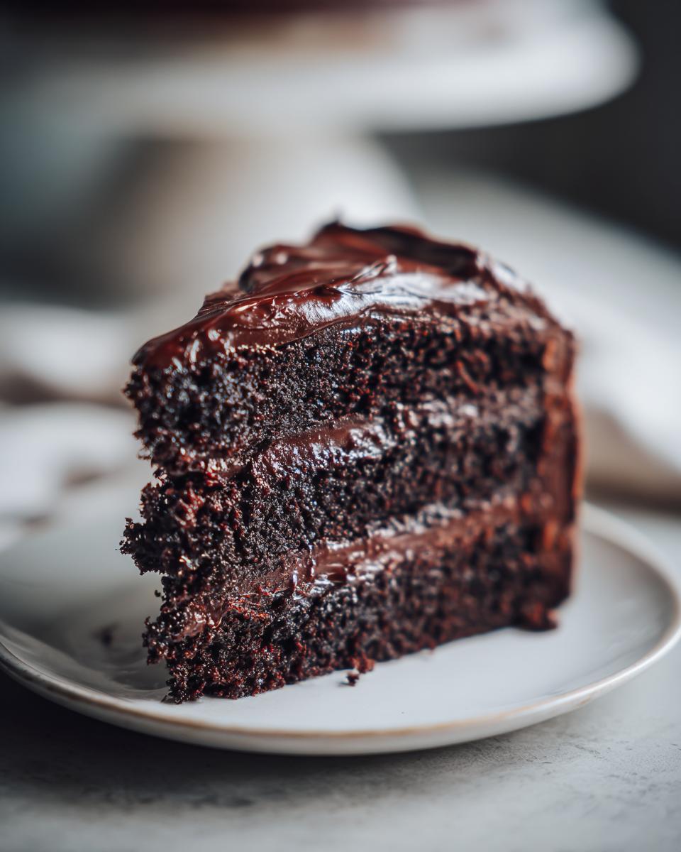 A decadent slice of three-layer chocolate cake covered in glossy chocolate frosting, served on a white plate.