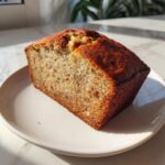 A thick slice of moist sugar free banana bread, featuring a golden crust and visible nuts, served on a white plate.